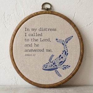 May include: A circular wooden embroidery hoop containing beige fabric with dark blue embroidery. The text reads: In my distress I called to the Lord, and he answered me. JONAH 2:2. Next to the text is a whale silhouette filled with floral patterns.