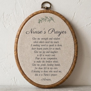 May include: An oval wooden embroidery hoop frame containing a beige fabric panel with the printed text Nurse's Prayer, a green leaf illustration, and a poem about nursing. The frame features a small metal hanging loop at the top center.