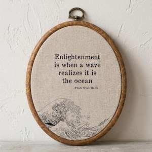May include: An oval embroidery hoop frame containing beige fabric with black stitched text that reads: Enlightenment is when a wave realizes it is the ocean -Thich Nhat Hanh. Below the text is a black stitched illustration of a large ocean wave.