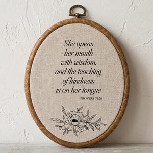 May include: An oval wooden embroidery hoop frame containing beige fabric with black printed text that reads: She opens her mouth with wisdom, and the teaching of kindness is on her tongue. PROVERBS 31:26. Below the text is a black floral line drawing.