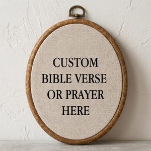 May include: An oval embroidery hoop frame with a wood grain finish containing light beige fabric. The fabric features the black printed text: CUSTOM BIBLE VERSE OR PRAYER HERE. A small metal hanging loop is attached to the top of the wooden frame.