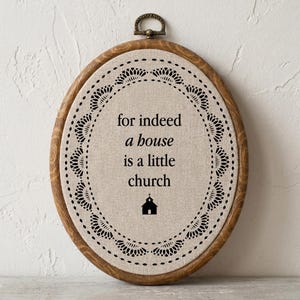 May include: An oval wooden embroidery hoop frame containing beige fabric with a black lace-style border. The text reads: for indeed a house is a little church, with a small black church icon centered below the text. A metal hanger is at the top.