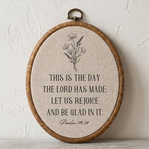 Peut inclure: Un cadre ovale en forme de tambour à broder en bois foncé contenant un tissu beige. Le tissu présente une illustration au trait noir de fleurs sauvages au-dessus du texte : THIS IS THE DAY THE LORD HAS MADE: LET US REJOICE AND BE GLAD IN IT. Psalm 118:24.