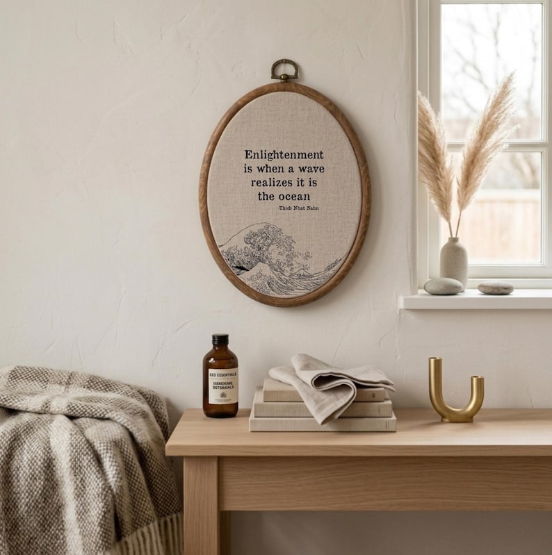May include: An oval wooden embroidery hoop wall hanging featuring beige fabric with black text that reads, "Enlightenment is when a wave realizes it is the ocean" by Thich Nhat Hanh, accompanied by a black line art illustration of a large ocean wave.