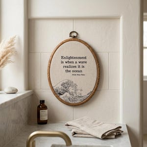 May include: An oval embroidery hoop wall hanging featuring a beige fabric background with a black line art illustration of a large ocean wave at the bottom and the text "Enlightenment is when a wave realizes it is the ocean" by Thich Nhat Nahn.