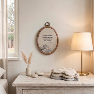 May include: An oval wooden embroidery hoop wall hanging featuring beige fabric with a black line art illustration of a wave and the text: Enlightenment is when a wave realizes it is the ocean, Thich Nhat Hanh, with a metal hanging loop at the top.