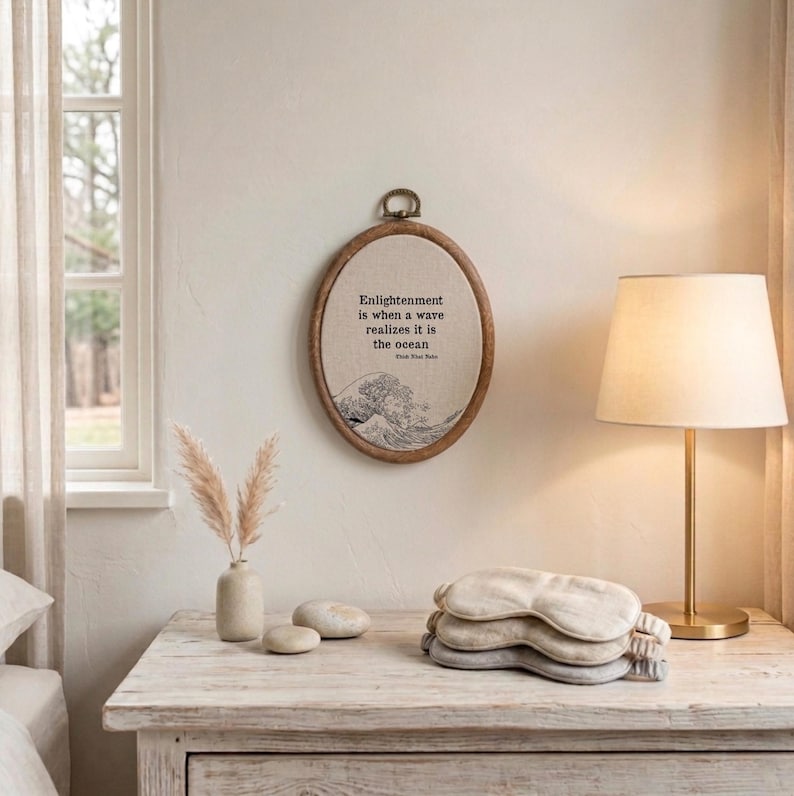 May include: An oval wooden embroidery hoop wall hanging featuring beige fabric with a black line art illustration of a wave and the text: Enlightenment is when a wave realizes it is the ocean, Thich Nhat Hanh, with a metal hanging loop at the top.