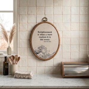 May include: An oval embroidery hoop wall hanging featuring a beige fabric background with a black line art illustration of a large ocean wave. The text reads: Enlightenment is when a wave realizes it is the ocean. Thich Nhat Nahn.