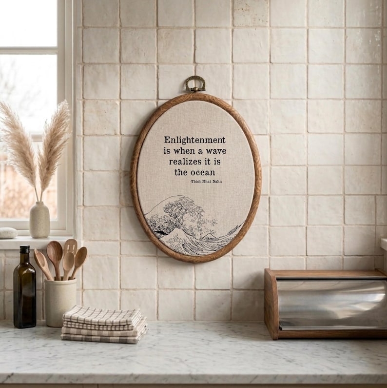May include: An oval embroidery hoop wall hanging featuring a beige fabric background with a black line art illustration of a large ocean wave. The text reads: Enlightenment is when a wave realizes it is the ocean. Thich Nhat Nahn.