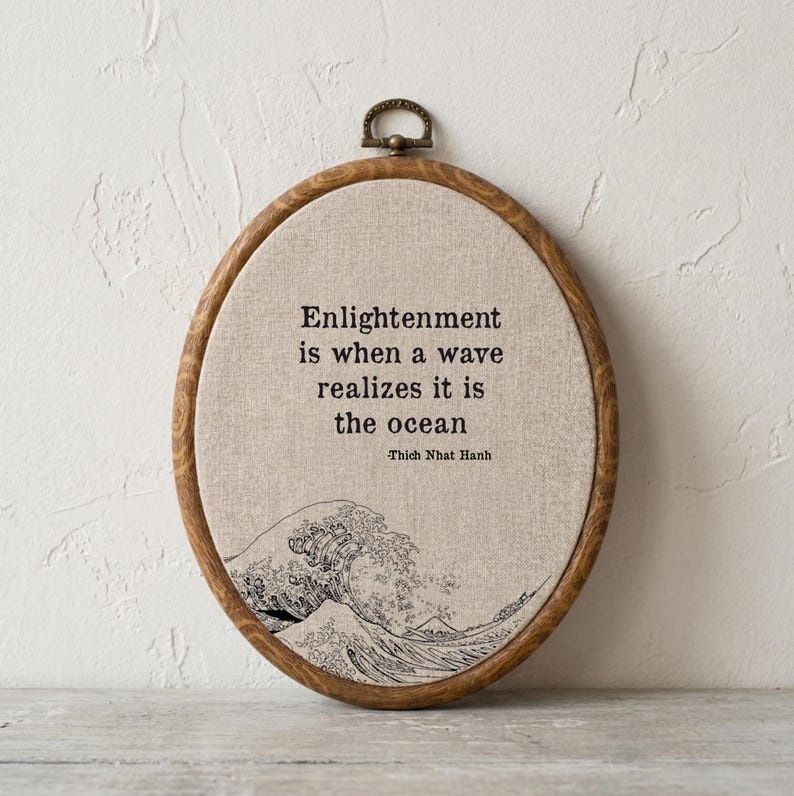 May include: An oval embroidery hoop frame containing beige fabric with black stitched text that reads: Enlightenment is when a wave realizes it is the ocean -Thich Nhat Hanh. Below the text is a black stitched illustration of a large ocean wave.