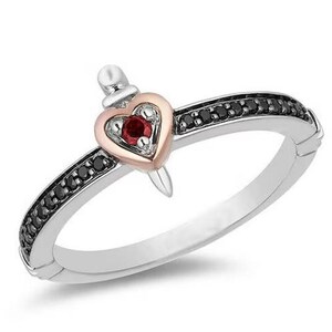 Op de afbeelding: Een zilveren ring met een centraal hart van roségoud dat doorboord wordt door een zilveren dolk. Het hart bevat een kleine rode edelsteen. De ringband is op de bovenste helft ingelegd met een rij kleine zwarte steentjes voor een contrast.