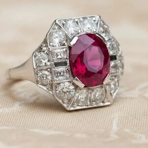 Può includere: Un anello color argento con un grande rubino rosso scuro a taglio ovale al centro. Il rubino è circondato da un alone geometrico di molteplici diamanti bianchi trasparenti a taglio rotondo e quadrato, incastonati in una montatura metallica.