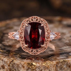 Può includere: Un anello in oro rosa con una grande gemma rosso scuro taglio cuscino al centro, circondata da una complessa cornice in metallo filigranato. Due pietre trasparenti più piccole taglio triangolare sono incastonate sui lati della fascia lavorata.
