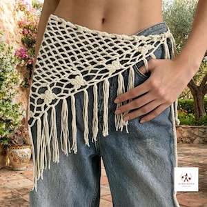 May include: An off-white crochet hip scarf featuring a diamond-shaped mesh pattern with small floral accents and long fringe trim, worn over blue denim jeans. The accessory is tied at the side and has a textured, knotted construction.