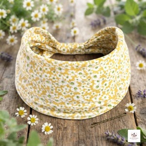 May include: A wide fabric headband featuring a mustard yellow background with a dense, repeating pattern of small white daisy flowers with green centres. The headband has a gathered, knotted detail at the top centre and a soft, flexible construction.