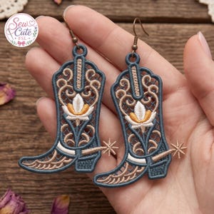 May include: A pair of dangling earrings shaped like cowboy boots, crafted from dark blue and tan thread with intricate lace-like embroidery. Each boot features a central white and gold floral motif and a small attached spur, suspended by bronze hooks.