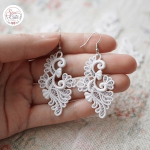May include: A pair of white lace earrings featuring intricate swirling patterns and floral-like motifs. The earrings are constructed from delicate thread work and hang from silver-toned metal hooks. A logo reading Sew Cute FSL is visible.