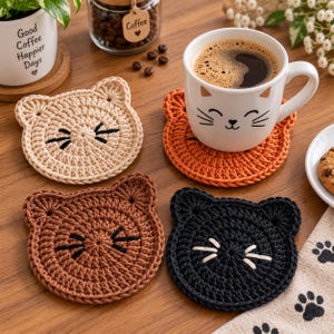 May include: Four round crochet coasters shaped like cat faces in beige, orange, brown, and black. Each coaster features embroidered whiskers and two small ears. One orange coaster supports a white ceramic mug with a cat face design.