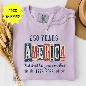 Puede incluir: Una camiseta de cuello redondo de color morado claro con el texto 250 YEARS AMERICA God shed his grace on thee 1776-2026. La palabra AMERICA está impresa en letras de bloque con patrones rojos, blancos y azules. Cuatro estrellas están bajo el texto.