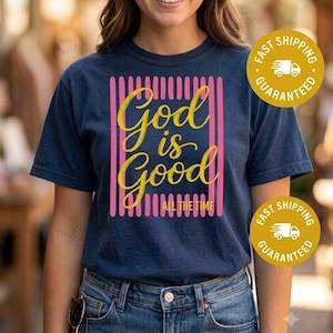 Puede incluir: Una camiseta azul marino de manga corta con un diseño gráfico de rayas verticales rosas y el texto amarillo en cursiva "God is Good" sobre el texto amarillo más pequeño "ALL THE TIME" centrado en la zona del pecho de la prenda.