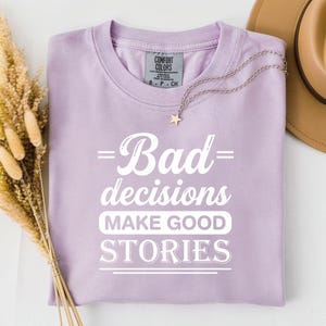 Puede incluir: Una camiseta de manga corta de color morado claro con texto blanco que dice "Bad decisions MAKE GOOD STORIES". La prenda cuenta con cuello redondo y una etiqueta de la marca Comfort Colors en el interior. Un collar con estrella plateada descansa sobre la tela.