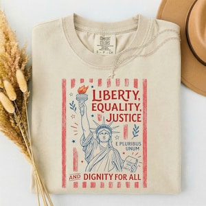 Puede incluir: Una camiseta beige de cuello redondo con un gráfico de la Estatua de la Libertad en tinta azul y roja. El texto dice: LIBERTY, EQUALITY, JUSTICE, E PLURIBUS UNUM, AND DIGNITY FOR ALL. El diseño está enmarcado por rayas rojas verticales.