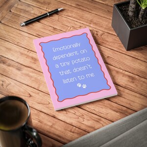 Puede incluir: Un cuaderno rectangular con cubierta rosa y un panel central azul claro delineado por un borde rojo ondulado. El texto dice: Emotionally dependent on a tiny potato that doesn't listen to me, acompañado por dos pequeñas huellas de pata blancas.