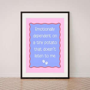 Könnte beinhalten: Ein gerahmter Kunstdruck mit einem hellblauen Rechteck mit gewelltem rotem Rand auf rosa Hintergrund. Der Text lautet: Emotionally dependent on a tiny potato that doesn't listen to me, darunter zwei kleine weiße Pfotenabdrücke.