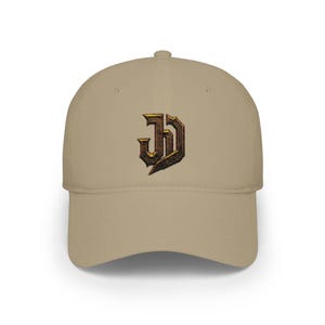 May include: A khaki baseball cap featuring a centred, embroidered logo with the letters J and D intertwined. The letters have a metallic, textured appearance with dark brown and gold tones, creating a sharp, angular, and gothic-inspired design.