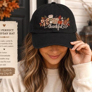 May include: A black baseball cap featuring a graphic print of a plaid bow, a coffee cup, a pumpkin, and a book with a cross, accompanied by the word thankful in white script. The cap has a curved brim and an adjustable strap at the back.