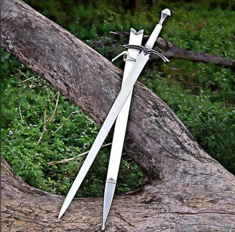 May include: A silver-toned metal sword with a pointed blade, a cross-shaped guard, and a white textured grip. The sword is accompanied by a matching white scabbard with silver metal fittings at the tip and throat, resting against a textured log.