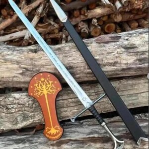 May include: A silver-toned metal sword with a black grip and a pommel, accompanied by a black scabbard with a silver tip. Beside the sword is a brown, shield-shaped plaque featuring a gold-coloured tree design with a crown at the top.