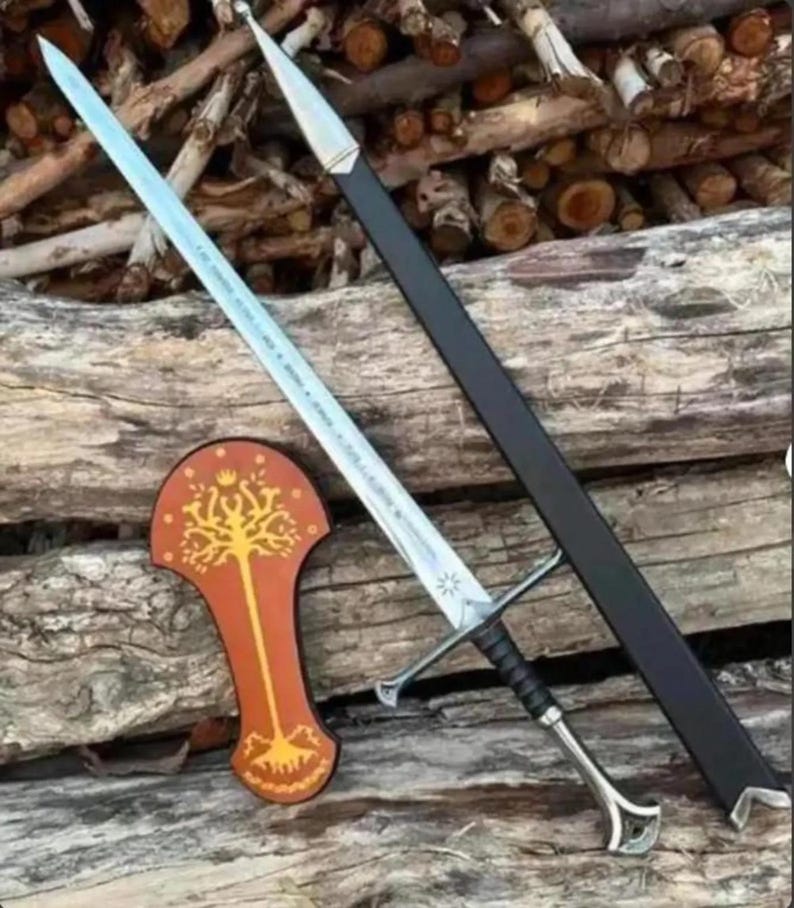 May include: A silver-toned metal sword with a black grip and a pommel, accompanied by a black scabbard with a silver tip. Beside the sword is a brown, shield-shaped plaque featuring a gold-coloured tree design with a crown at the top.