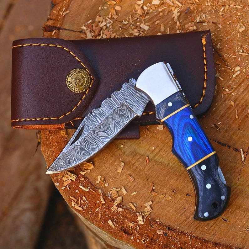 May include: A folding pocket knife with a Damascus steel blade featuring a wavy pattern, a silver bolster, and a handle with blue and black wood grain sections secured by silver pins. It is accompanied by a brown leather sheath with yellow stitching.