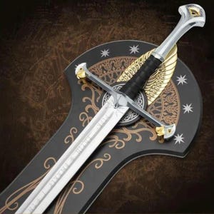 May include: A silver-toned sword with a black grip and gold-coloured accents on the crossguard and pommel. The sword is mounted on a black, shield-shaped plaque featuring gold filigree patterns, white stars, and a central golden wing design.