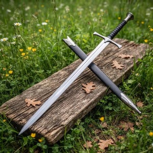 Puede incluir: Una espada de acero de doble filo con una guarda plateada y una empuñadura negra acanalada, acompañada de una vaina negra a juego con herrajes metálicos plateados en la punta y la boca, apoyada en diagonal sobre una tabla de madera desgastada.