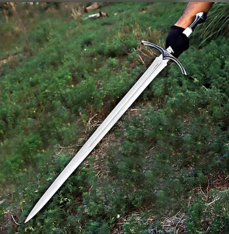 May include: A long, double-edged steel sword with a pointed tip and a central fuller running down the blade. The hilt features a curved crossguard with intricate silver-toned detailing and a black grip, held by a gloved hand against greenery.