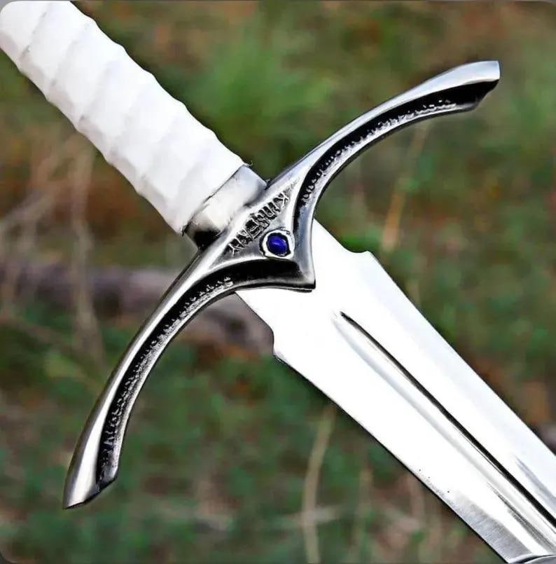 May include: A sword featuring a polished silver-tone metal crossguard with an engraved inscription and a central blue gemstone. The handle is wrapped in a textured white material, and the blade is made of smooth, reflective silver-tone metal.