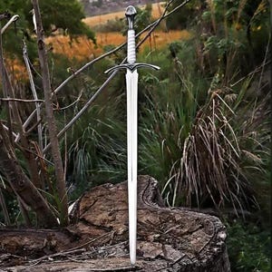 May include: A silver-toned metal sword featuring a long, pointed blade, a crossguard with decorative detailing, and a white wrapped handle topped with a rounded pommel. The sword is positioned vertically, with its tip resting on a wooden surface.