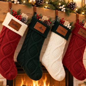 May include: Four cable-knit Christmas stockings in red, dark green, and white, each featuring a leather-like name tag labelled Jordan, Alex, Ethan, or Morgan. The stockings are filled with small pinecones, red berries, and wrapped gift boxes.