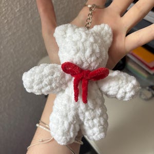 Peut inclure: Un porte-clés en forme d'ours en peluche blanc, réalisé en fil texturé doux. L'ours est orné d'un petit nœud rouge vif en velours autour du cou et est fixé à un anneau fendu en métal argenté avec une courte chaînette pour l'accrocher.