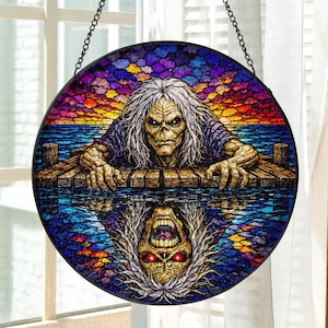 May include: A circular stained glass suncatcher featuring a long-haired figure with a skeletal face resting on a wooden dock. The scene includes a vibrant sunset sky, blue water, and a reflection of the figure with glowing red eyes below.