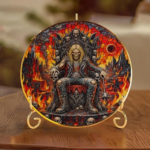 Può includere: Un piatto decorativo circolare con un'illustrazione in stile vetrata raffigurante una figura scheletrica dai capelli lunghi con giacca di pelle seduta su un trono circondato da teschi, fiamme e un paesaggio oscuro, su un supporto in metallo dorato.