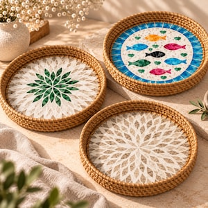 May include: Three round woven rattan trays with raised edges. Two trays feature a white and green floral-like petal pattern. The third tray displays a central white circle with colorful fish in orange, blue, pink, and black, surrounded by a blue border.