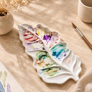 Può includere: Una tavolozza per pittura in ceramica bianca a forma di foglia con scomparti multipli contenenti vari pigmenti per acquerello essiccati nelle tonalità del rosso, viola, giallo, verde e blu, con un design a venatura centrale.