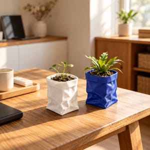 May include: Two square plant pots with a crinkled paper-like texture, one in solid white and one in solid blue, each containing a small green plant with soil visible at the top, positioned on a wooden surface in a bright indoor setting.
