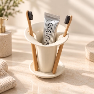 May include: A cream-colored ceramic toothbrush holder with a wide, flared rim and a circular base. The holder contains two bamboo toothbrushes with black bristles and a silver tube of Marvis toothpaste with black text printed on the front.