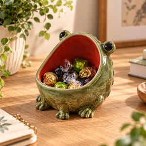 May include: A ceramic frog-shaped bowl with a mottled green exterior and a smooth, vibrant red interior. The bowl features two prominent black eyes on top and small sculpted feet at the base, currently filled with several individually wrapped sweets.