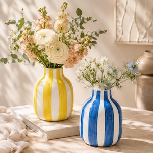 May include: Two ceramic vases with rounded bodies and narrow necks. One vase features vertical yellow and white stripes, filled with a large bouquet of pink, white, and cream flowers. The other vase has vertical blue and white stripes with small white and blue flowers.