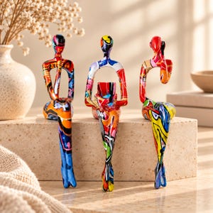 Puede incluir: Tres figuras abstractas con forma humana en posición sentada, cada una con un vibrante patrón de grafiti multicolor con llamativas salpicaduras de pintura roja, azul, amarilla y negra que cubren sus superficies lisas y estilizadas.